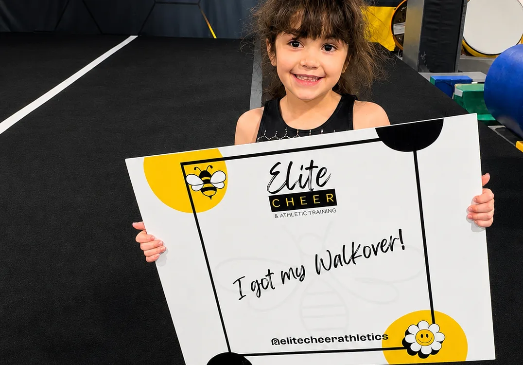 Young Elite Cheer athlete smiling with an 'I got my Walkover!' milestone sign in the gym